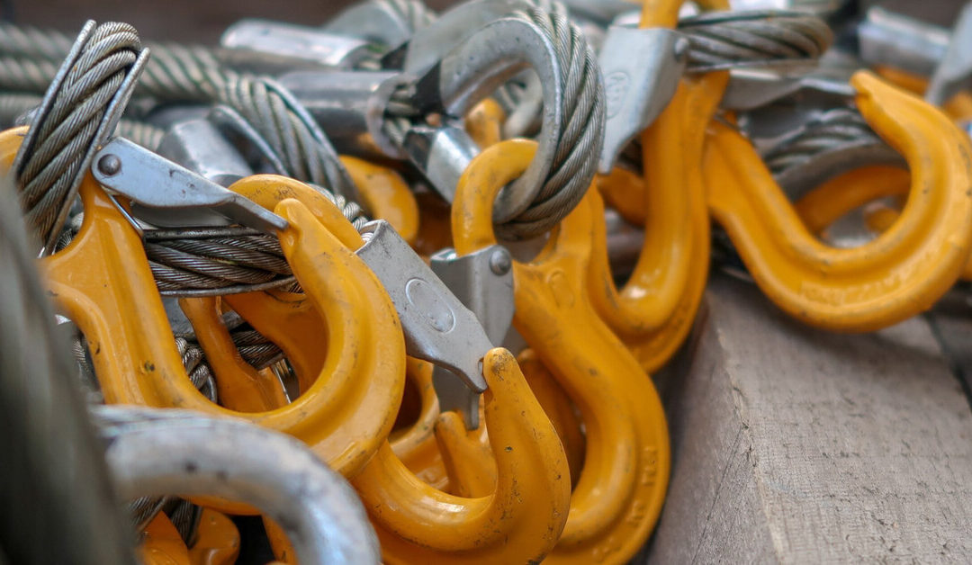 A large pile of bright yellow-colored sling hooks, each securely attached to thick, coiled wire ropes, rests on a wooden plank.