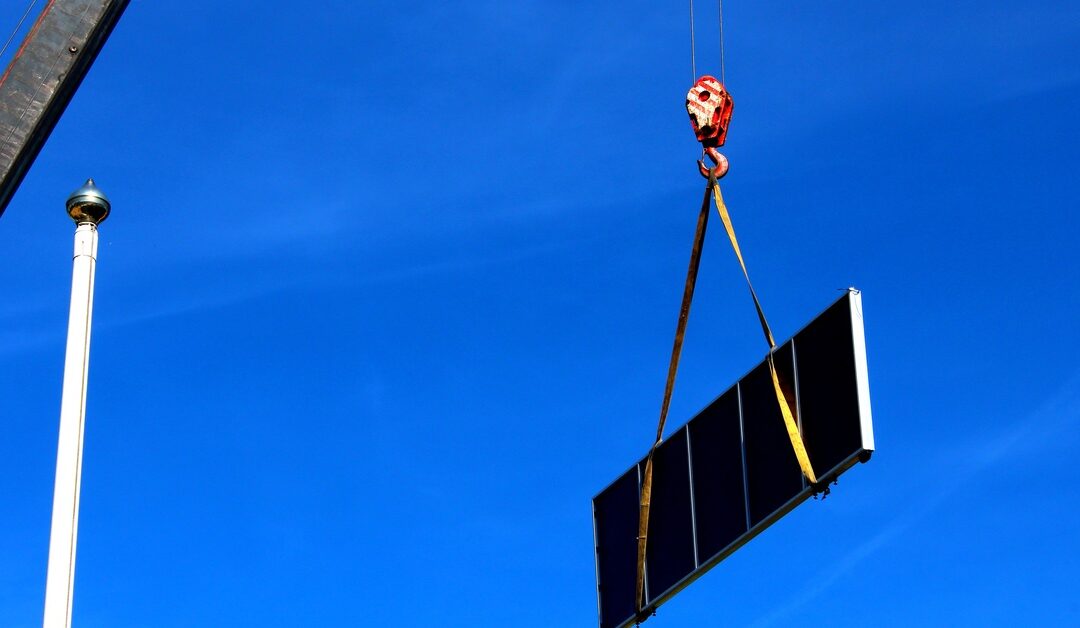 A crane lifts a large solar panel with slings and a hook against a clear blue sky during installation work.