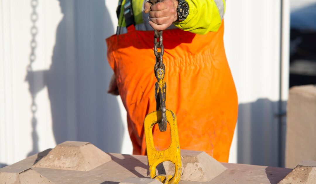 A worker in high-visibility gear holds a chain hook attached to a concrete slab at an outdoor jobsite.